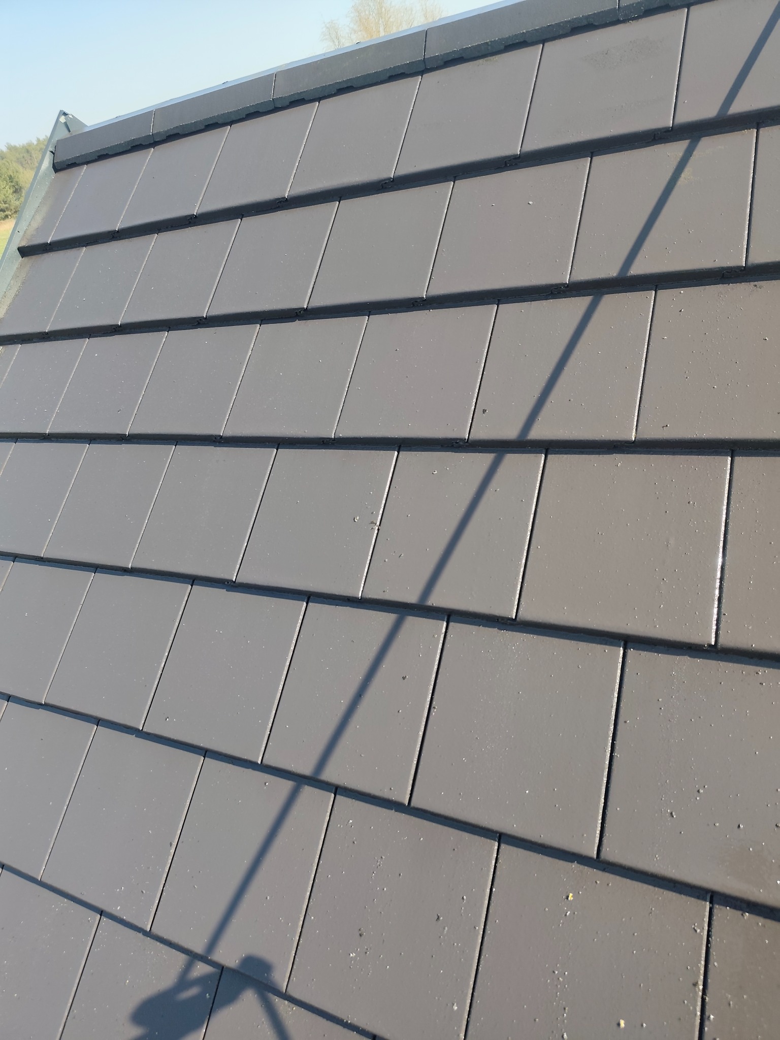 Czysty, szary dach z ceramicznej dachówki w słoneczny dzień. Widoczny cień osoby w lewym dolnym rogu. Dach po renowacji lub świeżo położony.