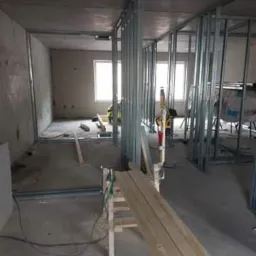 Wnętrze w trakcie montażu metalowej konstrukcji pod płyty gipsowo-kartonowe, widoczne profile, drewniane deski na kozłach, narzędzia w tle, surowy beton na podłodze i suficie.