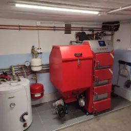 Czerwony piec CO marki Stalmark z podajnikiem, biały zasobnik ciepłej wody użytkowej i miedziana instalacja grzewcza w kotłowni z szarą podłogą.