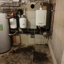 Biały piec centralnego ogrzewania z podłączeniami na ścianie z płytek, obok zbiornik wody i dwa zbiorniki wyrównawcze, widoczne rury i instalacje w piwnicy.