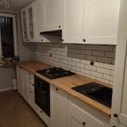 Nowoczesna kuchnia z białymi szafkami, drewnianym blatem, czarnym zlewem i płytą grzewczą, wykończona białymi płytkami z ciemną fugą.