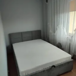 Szare łóżko tapicerowane z zagłówkiem w kształcie paneli, biały materac, jasne zasłony w tle, widoczna kratka wentylacyjna na ścianie.