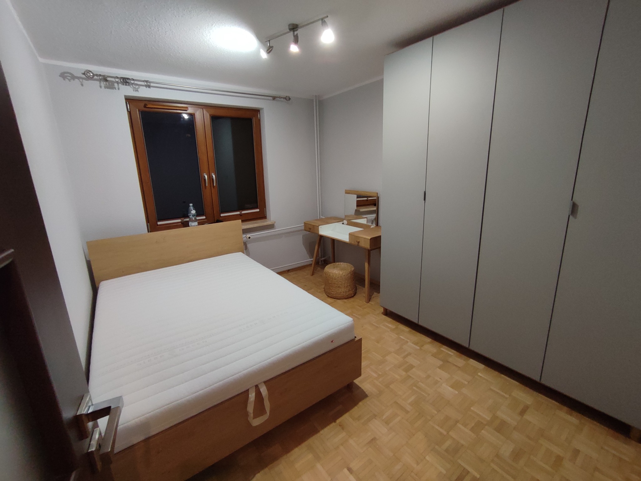 Jasna sypialnia z drewnianym łóżkiem, toaletką z lustrem i dużą, szarą szafą przesuwną na drewnianej podłodze.