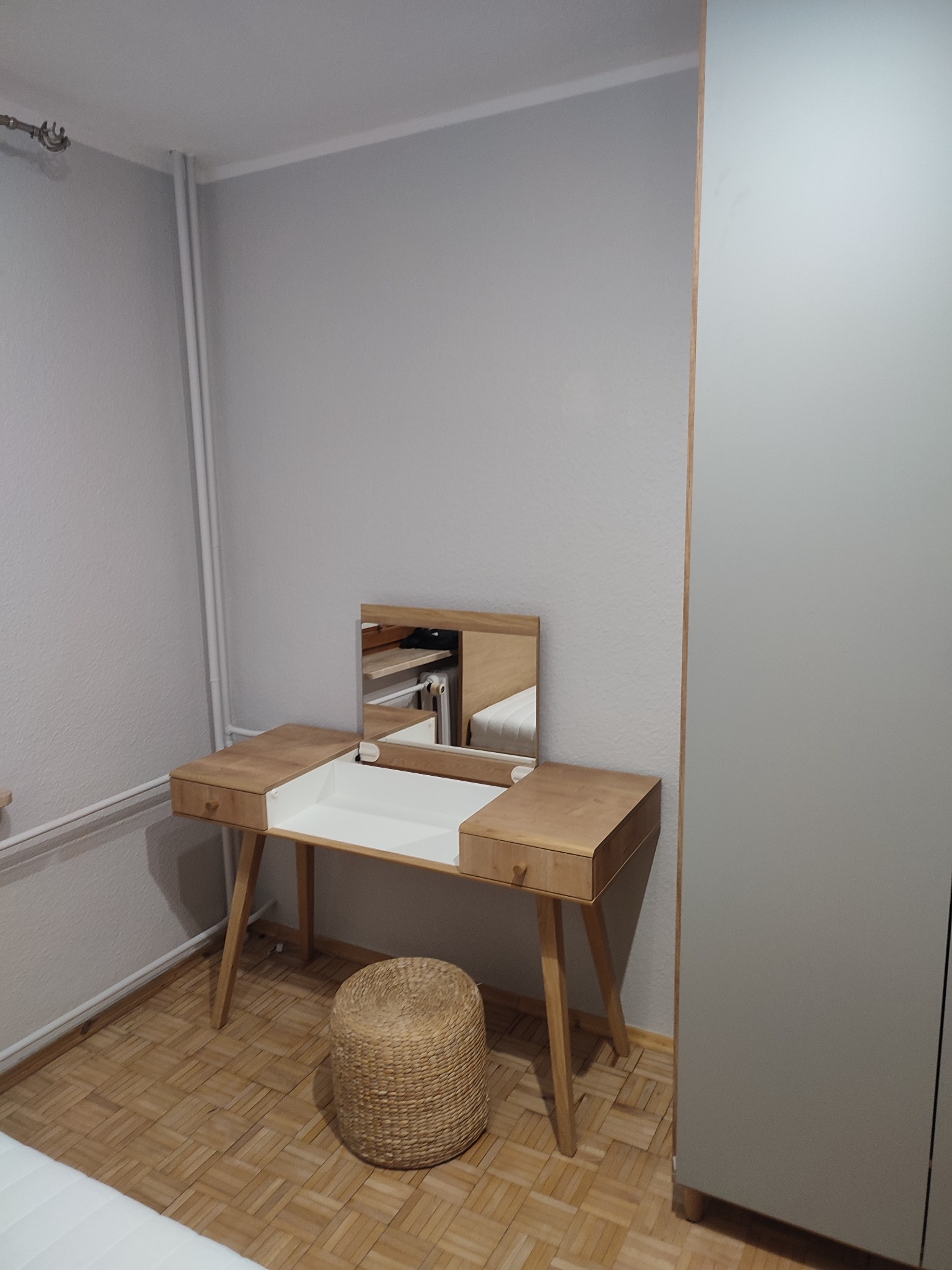 Drewniana toaletka z podnoszonym lustrem i wysuwanymi szufladami, ustawiona obok szarej szafy w pokoju z parkietem i wiklinowym pufem.