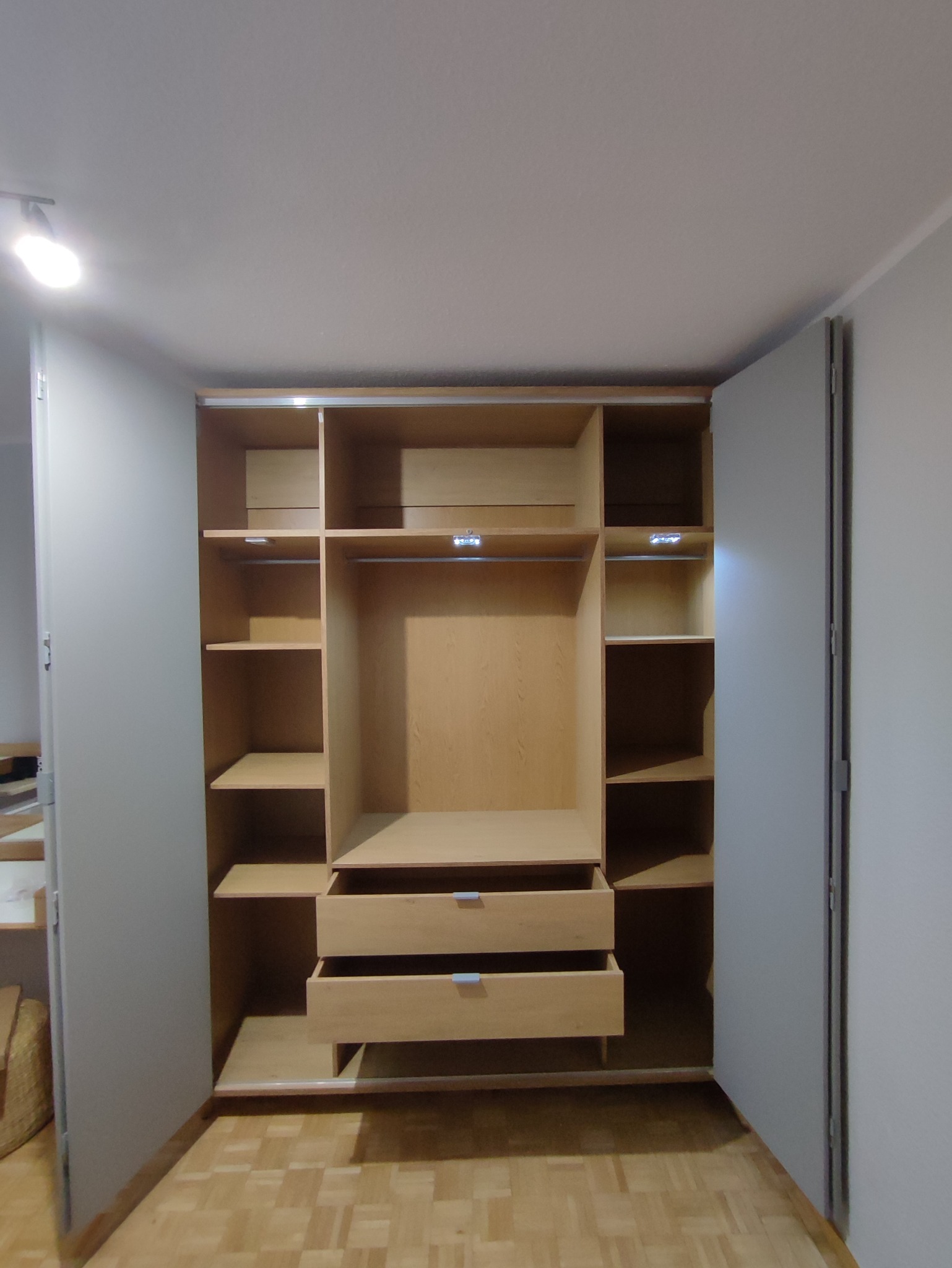 Otwarta szafa z jasnego drewna z półkami i dwiema wysuniętymi szufladami, widoczne oświetlenie LED wewnątrz, na podłodze parkiet.