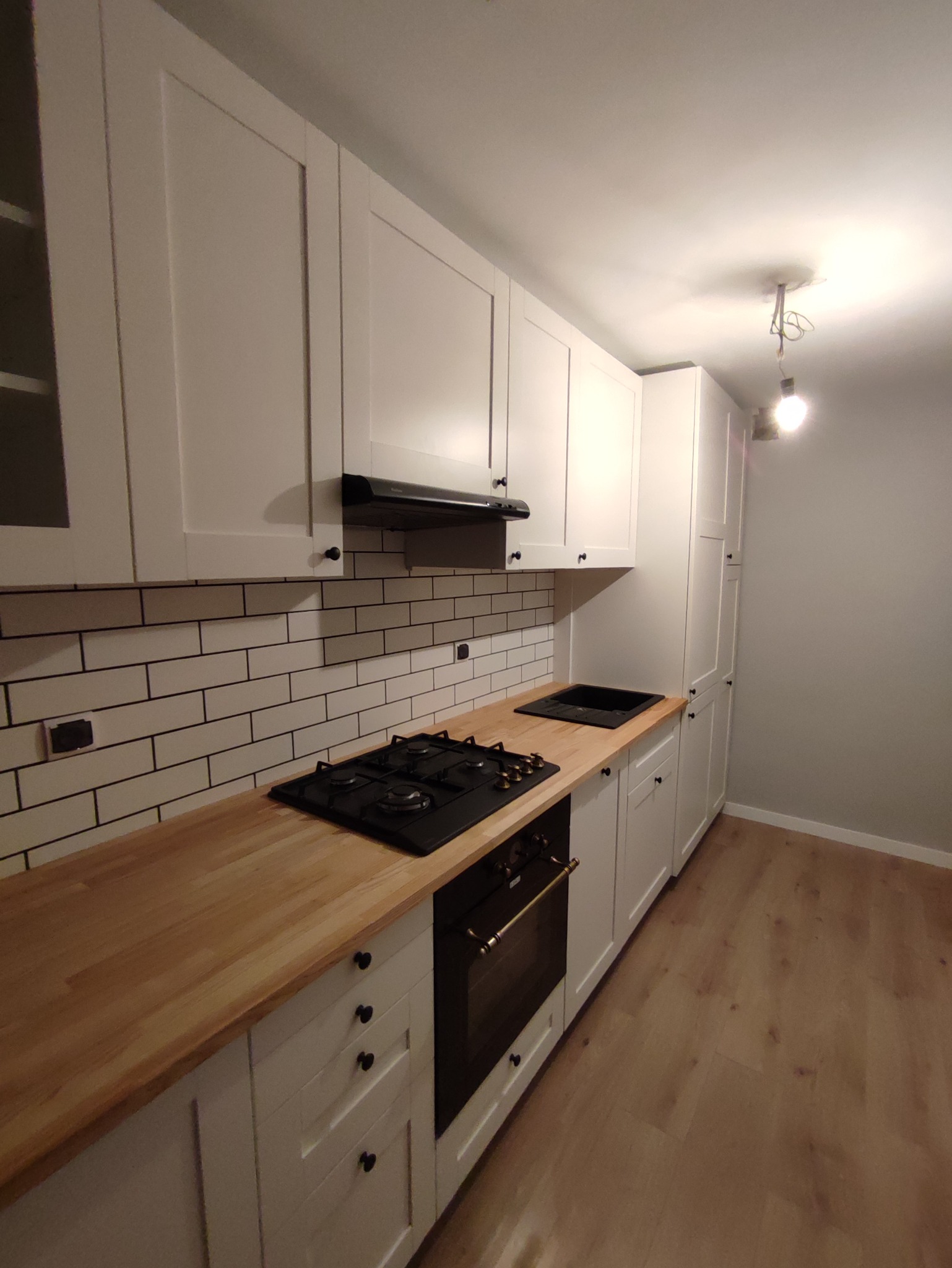 Nowoczesna kuchnia z białymi szafkami, drewnianym blatem, czarną płytą gazową i piekarnikiem w zabudowie, wykończona białymi płytkami z czarną fugą.