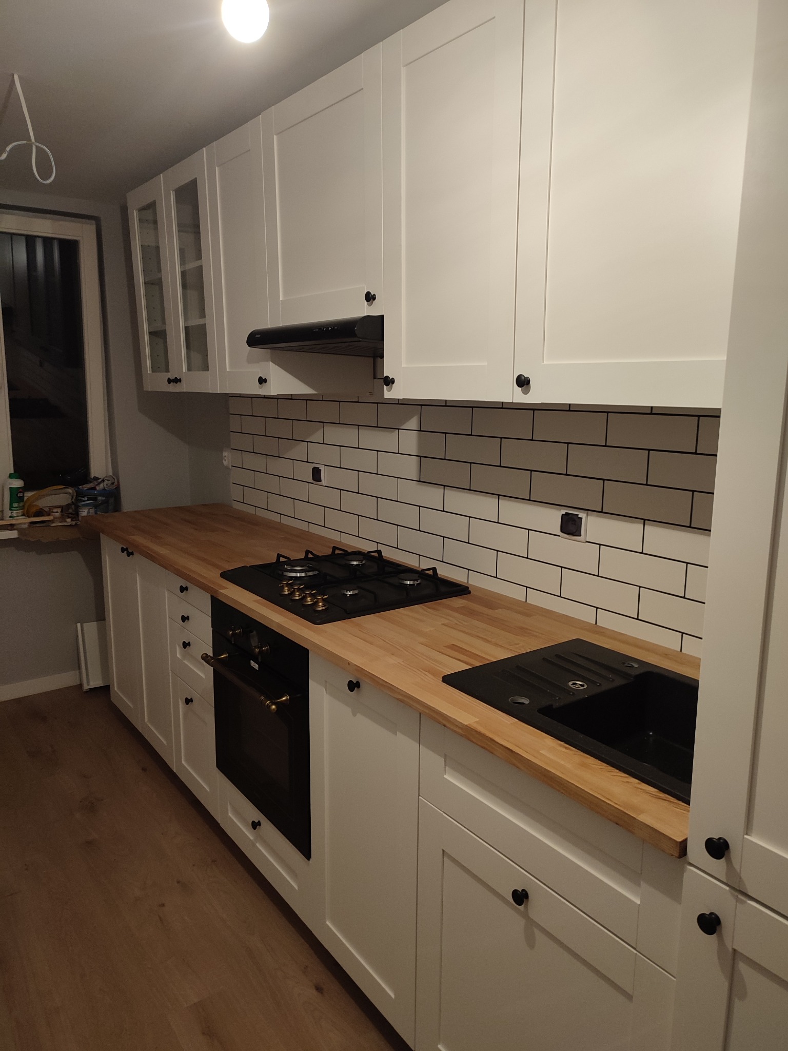 Nowoczesna kuchnia z białymi szafkami, drewnianym blatem, czarnym zlewem i płytą grzewczą, wykończona białymi płytkami z ciemną fugą.