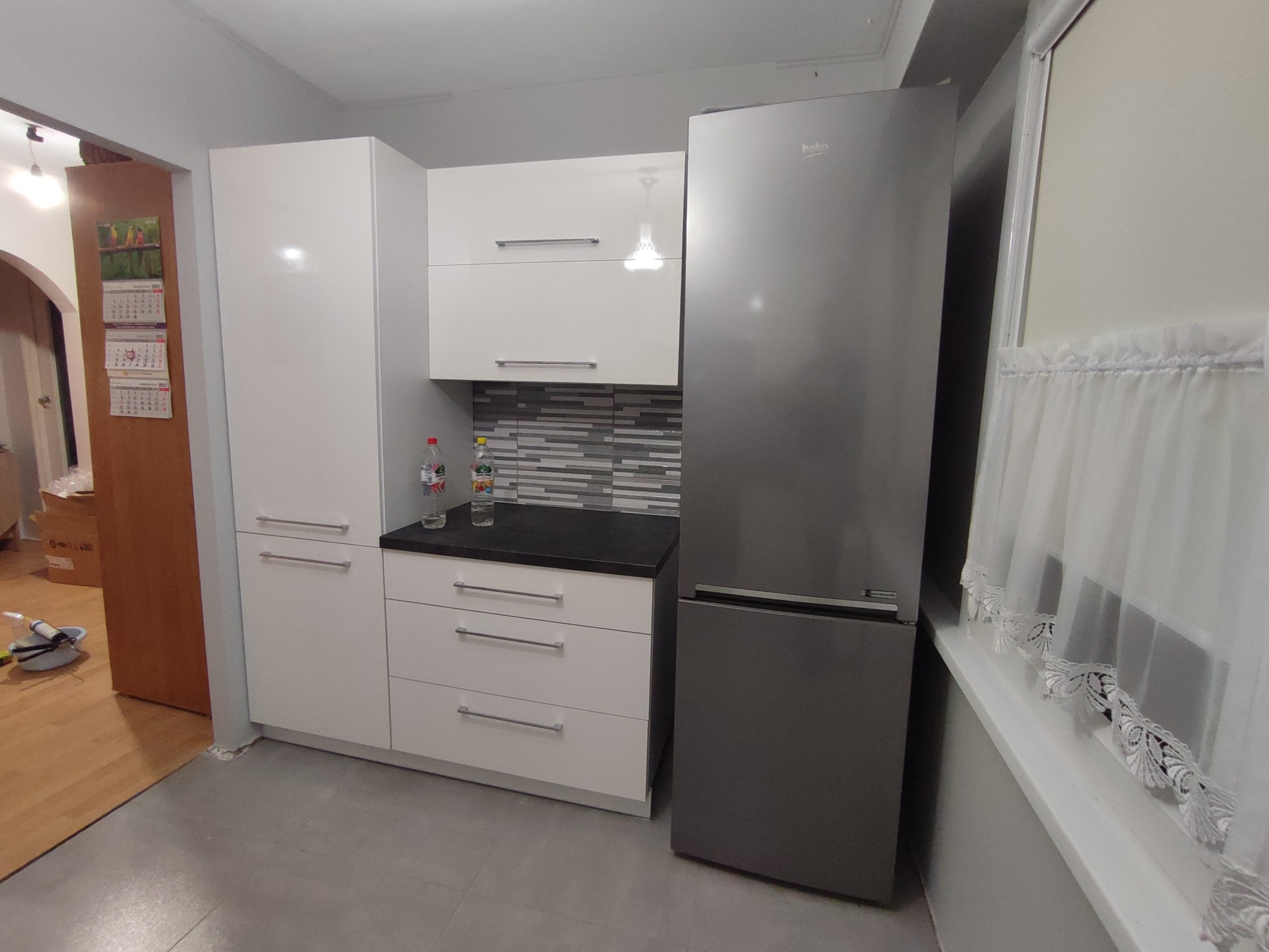 Nowoczesna kuchnia z białymi szafkami, czarnym blatem, szarymi płytkami i srebrną lodówką Beko, uzupełniona o białe, koronkowe firanki w oknie.