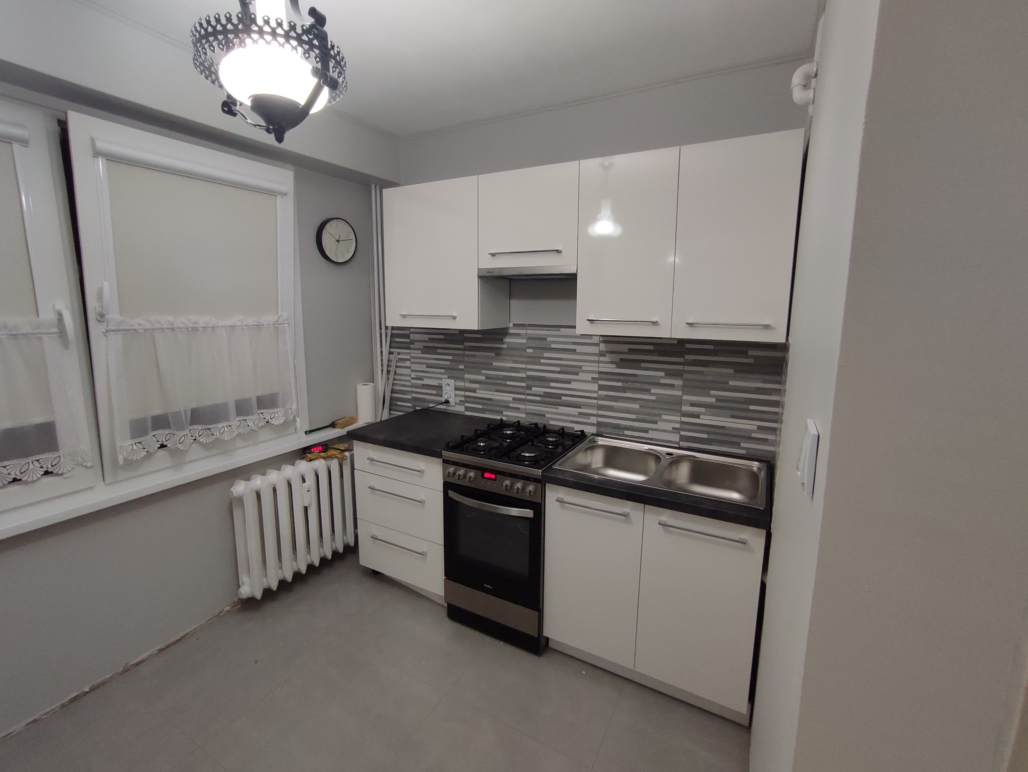 Nowoczesna kuchnia z białymi, błyszczącymi szafkami, szarym blatem, dwukomorowym zlewem i płytą gazową z piekarnikiem, z dekoracyjnym oświetleniem sufitowym i szaro-białą mozaiką na ścianie.