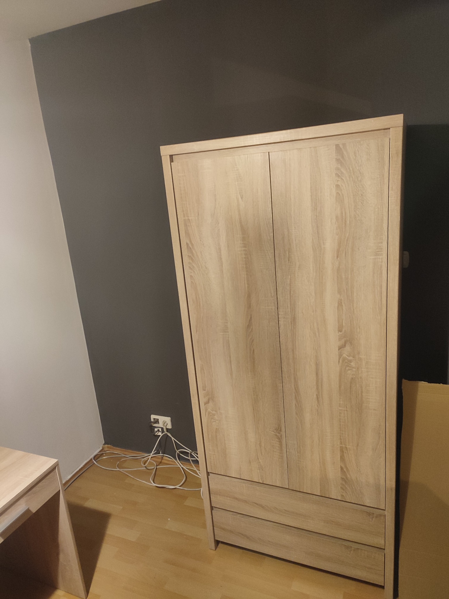 Dwudrzwiowa szafa z jasnego drewna z dwiema szufladami u dołu, ustawiona na drewnianej podłodze przy ścianie pomalowanej w dwóch odcieniach szarości, widoczne gniazdko elektryczne i plątanina kabli.