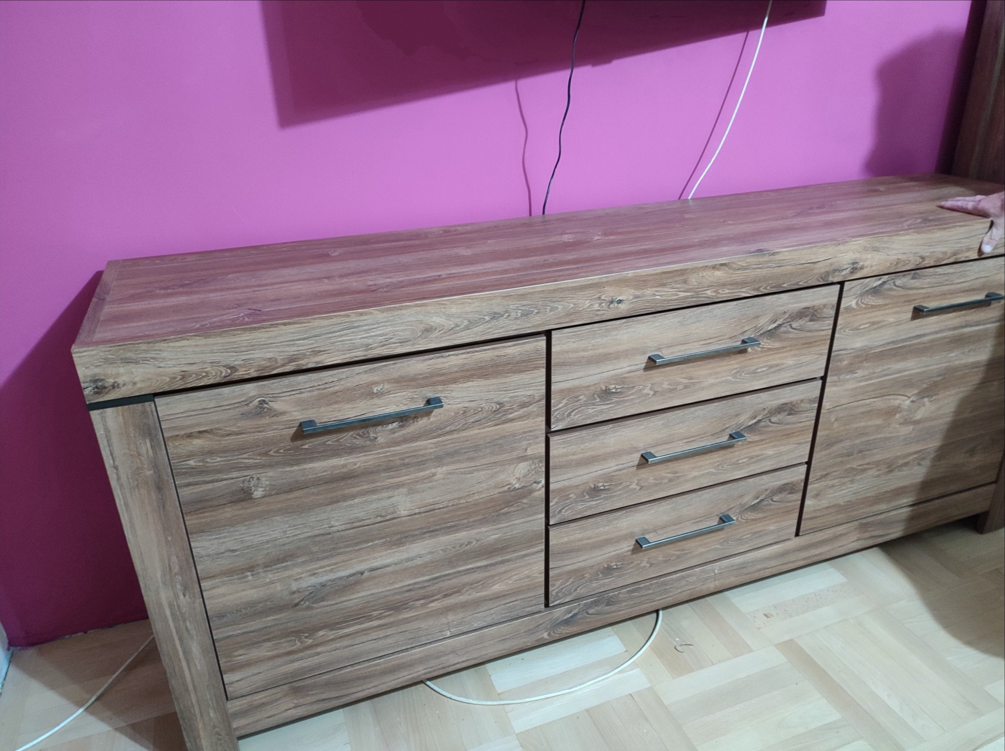 Komoda RTV z drewnopodobnym fornirem i metalowymi uchwytami na tle fuksjowej ściany, widoczne kable z tyłu.