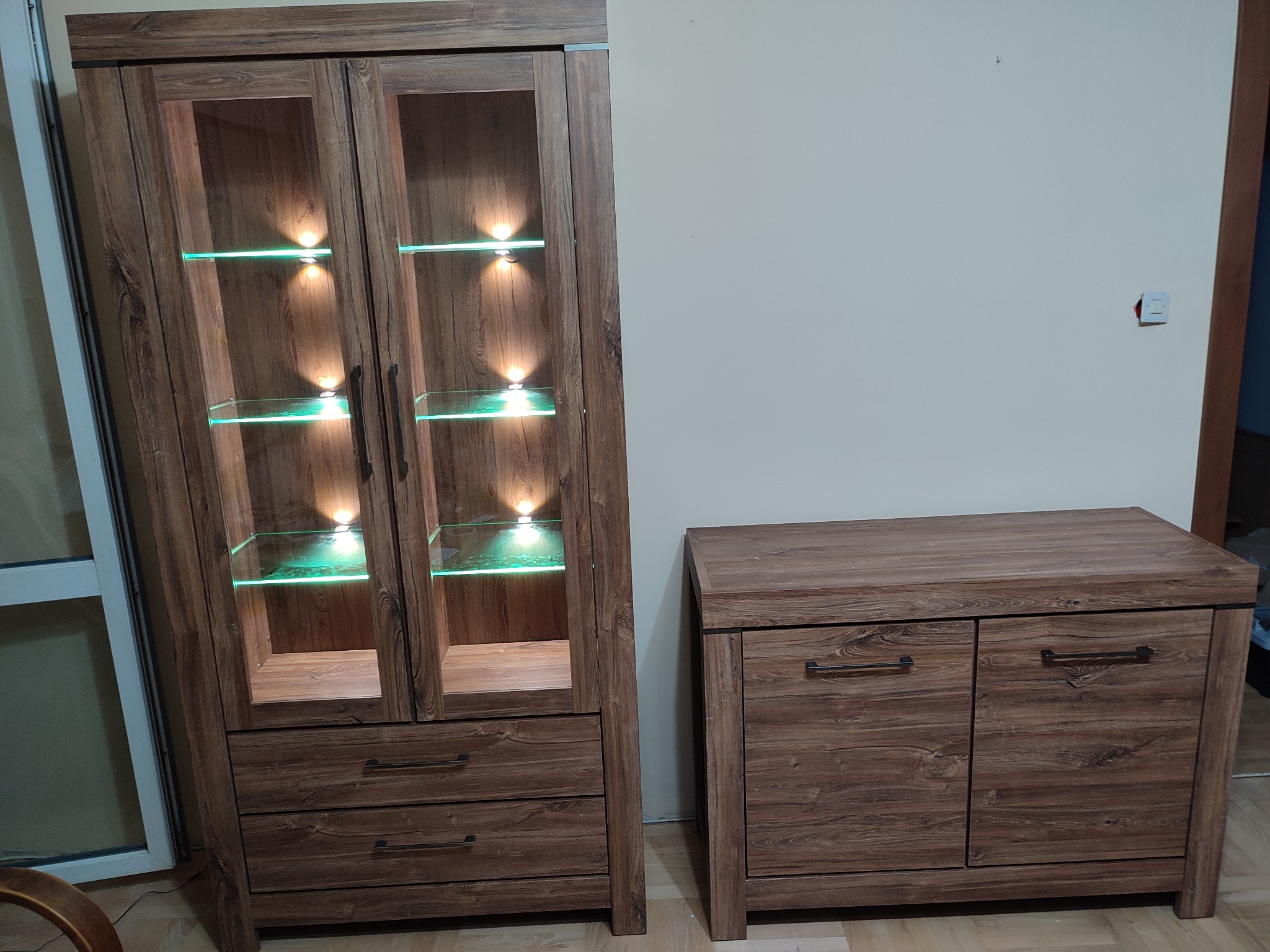 Zestaw mebli do salonu z forniru drewnopodobnego: witryna z przeszklonymi drzwiczkami i podświetlanymi półkami oraz komoda z dwoma szafkami, uchwyty w kolorze czarnym.