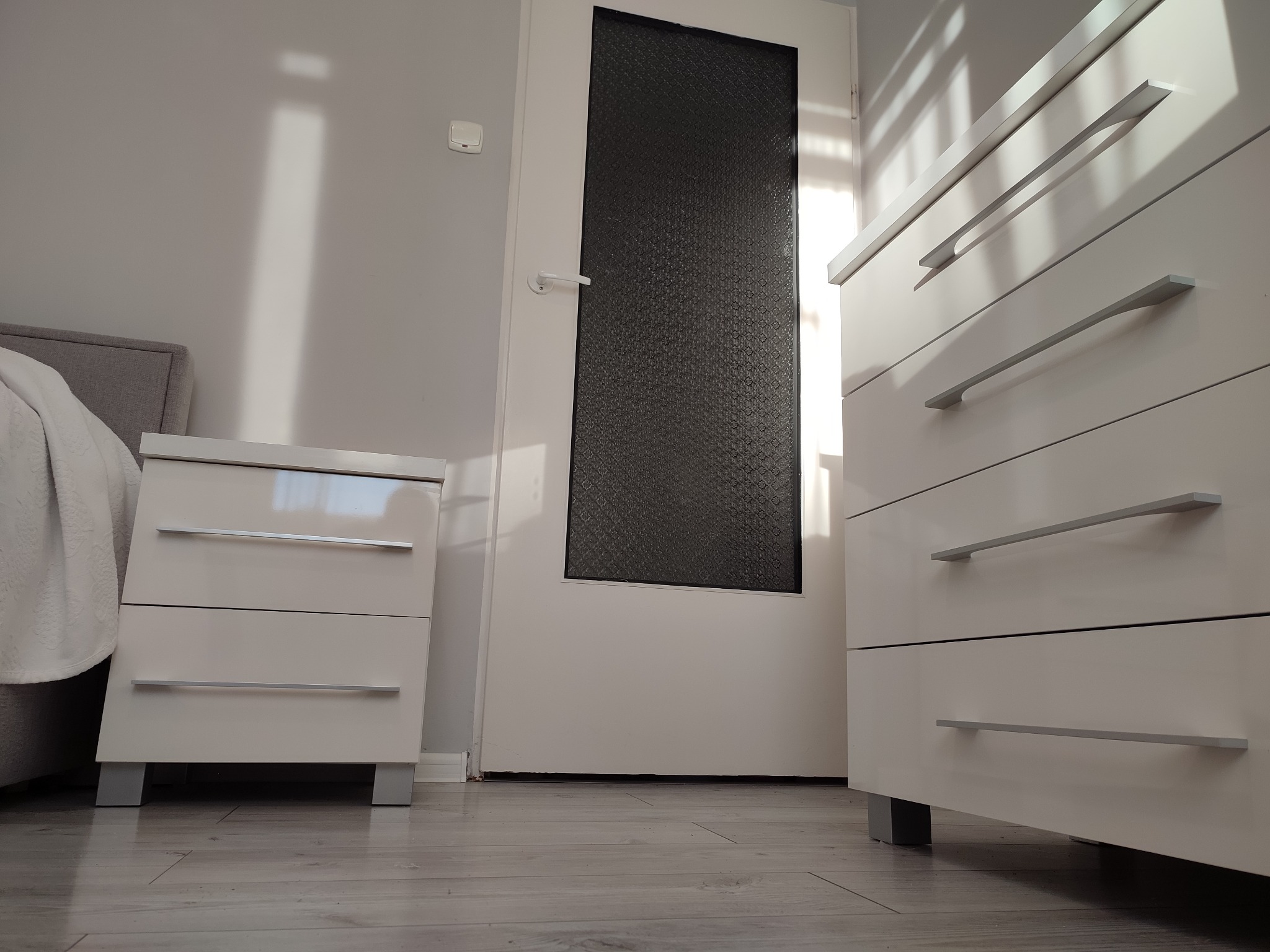 Nowoczesna, biała szafka nocna i komoda z podłużnymi, srebrnymi uchwytami, ustawione w sypialni w Tarnobrzegu, obok łóżka i drzwi z czarnym, zdobionym szkłem.