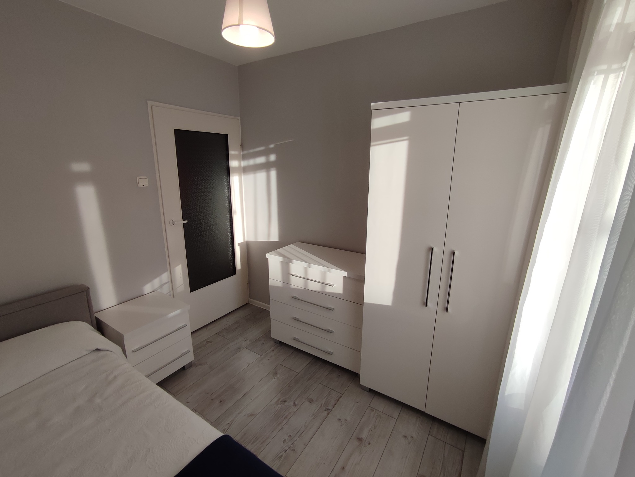 Jasne wnętrze sypialni z białymi meblami: szafa, komoda i szafka nocna, oświetlone popołudniowym słońcem wpadającym przez okno z delikatną firanką.