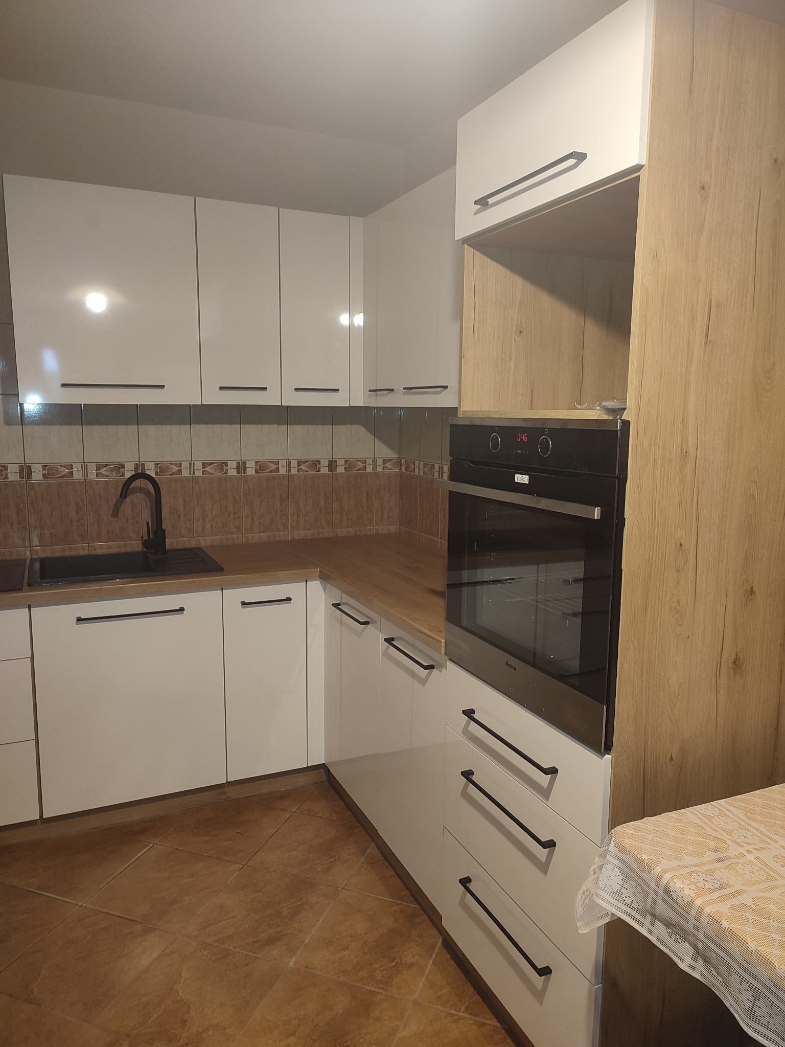 Nowoczesna kuchnia z białymi szafkami, czarną armaturą i piekarnikiem w zabudowie, z blatem imitującym drewno i brązowymi płytkami na ścianie.