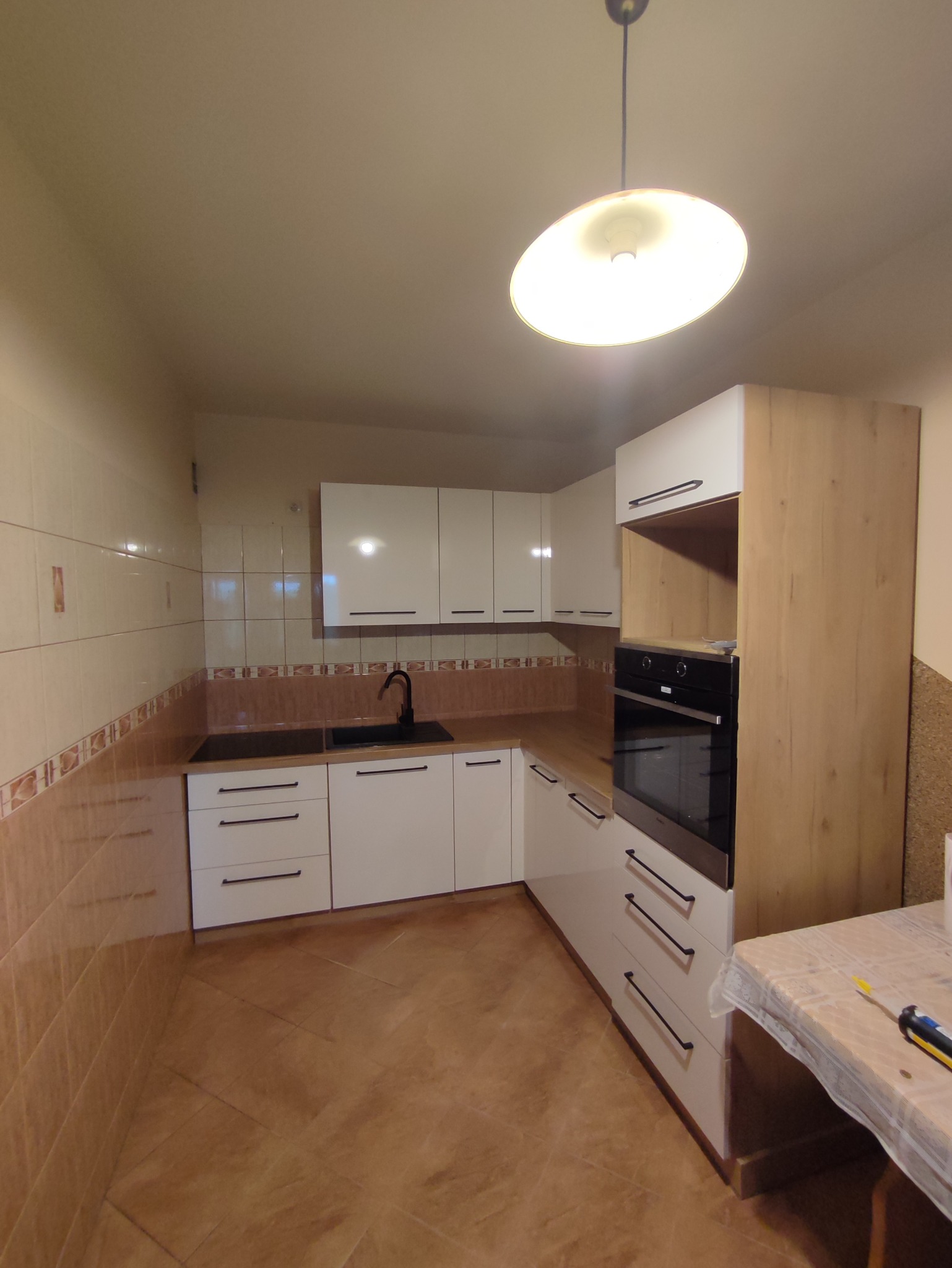 Nowoczesna kuchnia z białymi frontami szafek i drewnopodobnym wykończeniem, czarną armaturą i piekarnikiem w zabudowie, widok na fragment blatu i narzędzia na stole.