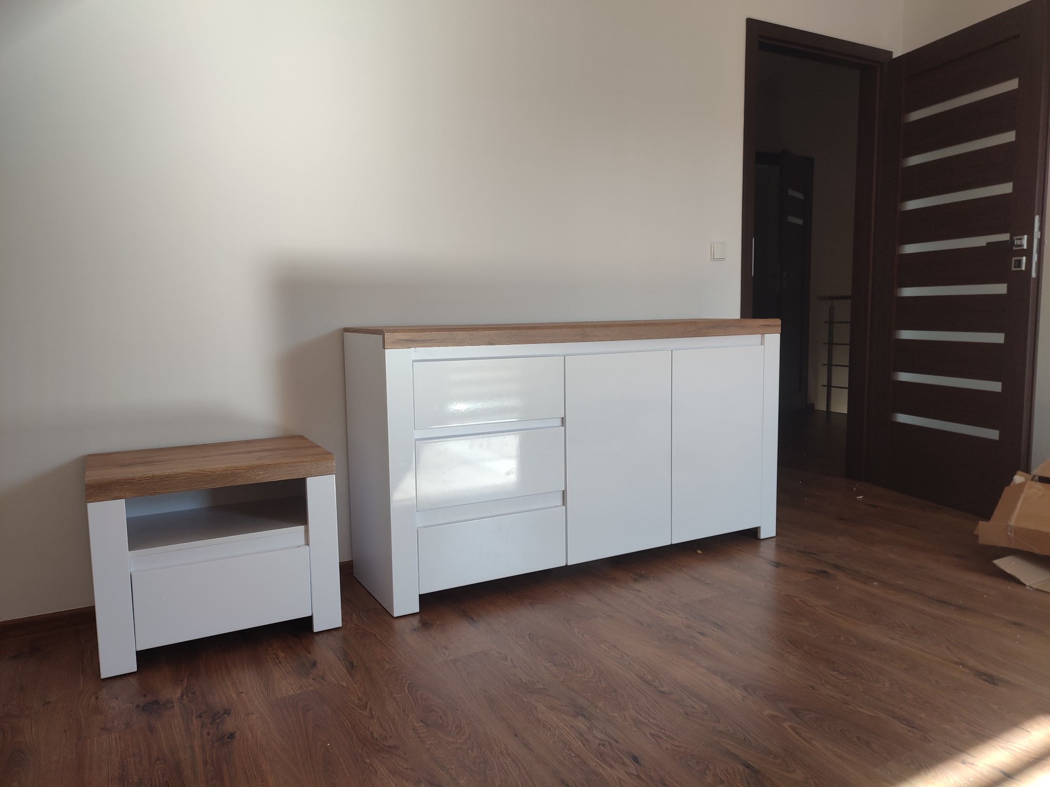 Nowoczesna biała komoda z drewnianym blatem i szafka nocna z tej samej serii, ustawione na brązowej podłodze, z widocznymi elementami kartonu w tle i otwartymi drzwiami do innego pomieszczenia.