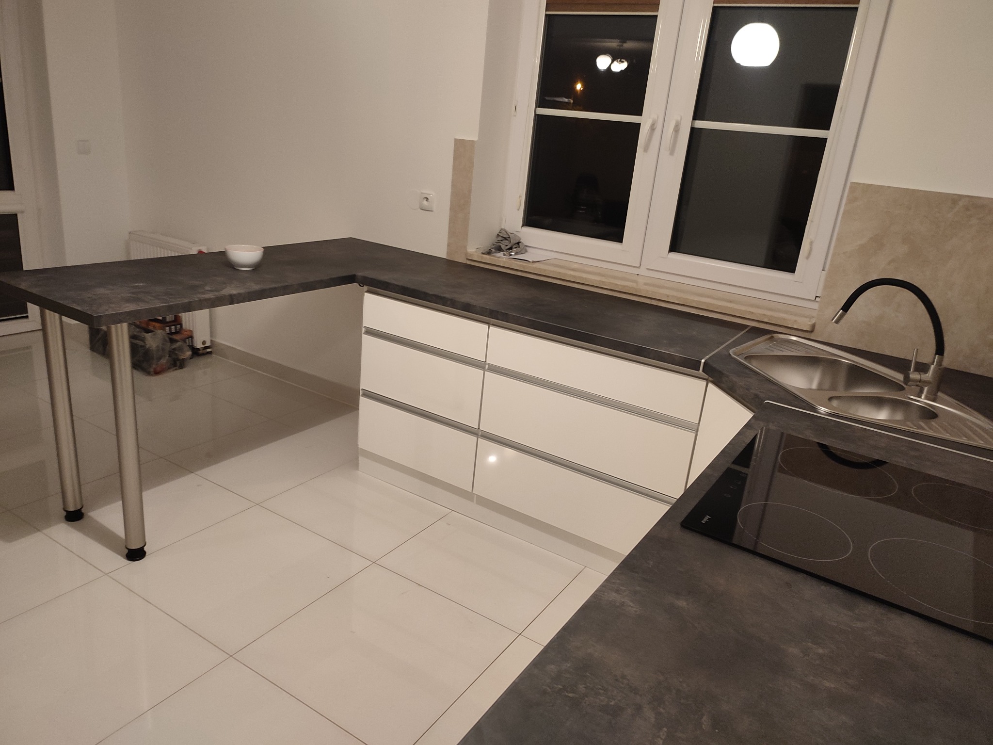 Nowoczesna kuchnia z szarym blatem, białymi frontami szafek, płytą indukcyjną i zlewem pod oknem, uzupełniona o stół na metalowych nogach z białą miseczką.