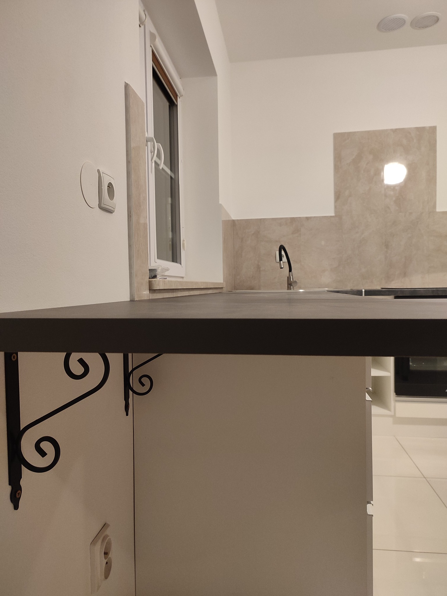 Czarny blat kuchenny wsparty na ozdobnych, kutych wspornikach w nowocześnie wykończonej kuchni z jasnymi płytkami i oknem.