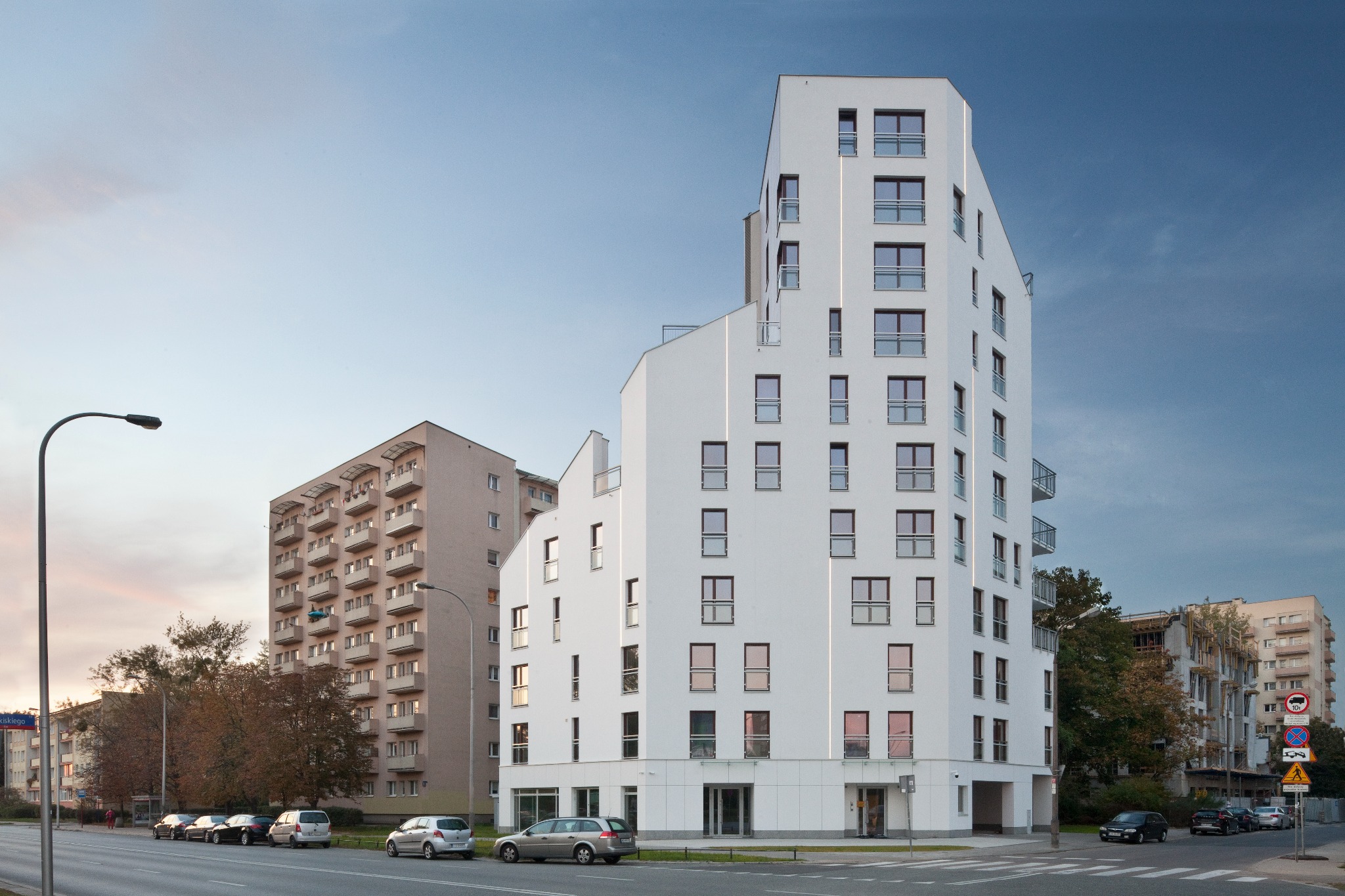 Nowoczesny, biały budynek apartamentowy z balkonami w Warszawie, widok z ulicy Woronicza przy skrzyżowaniu, w tle starszy blok mieszkalny i drzewa.