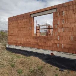 Wznoszenie ściany z czerwonej ceramiki poryzowanej, widoczny otwór okienny z nadprożem, izolacja pionowa fundamentu z folii kubełkowej, w tle rusztowanie.