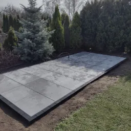 Ułożony taras z szarych płyt na gruncie, obramowany ciemnymi kostkami, otoczony zielenią i drzewami iglastymi.