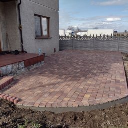 Przedsiębiorstwo Budowlane Waldemar Szewczyk - Nowo ułożony, brązowo-pomarańczowy chodnik z kostki brukowej przed domem, z obrzeżem z małych, okrągłych kostek, widoczne schody wejściowe z tego samego materiału i szara betonowa ściana budynku...