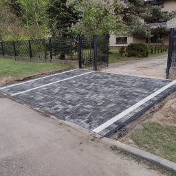 Przedsiębiorstwo Budowlane Waldemar Szewczyk - Nowo ułożony podjazd z szaro-czarnej kostki brukowej z białymi liniami, prowadzący do metalowej bramy posesji z domem w tle, widoczny fragment ulicy i trawnika z kupką piasku.