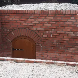 Wejście do piwniczki ziemnej z murowaną fasadą z czerwonej cegły, z drewnianymi drzwiami łukowymi, otoczone ziemią pokrytą warstwą śniegu.