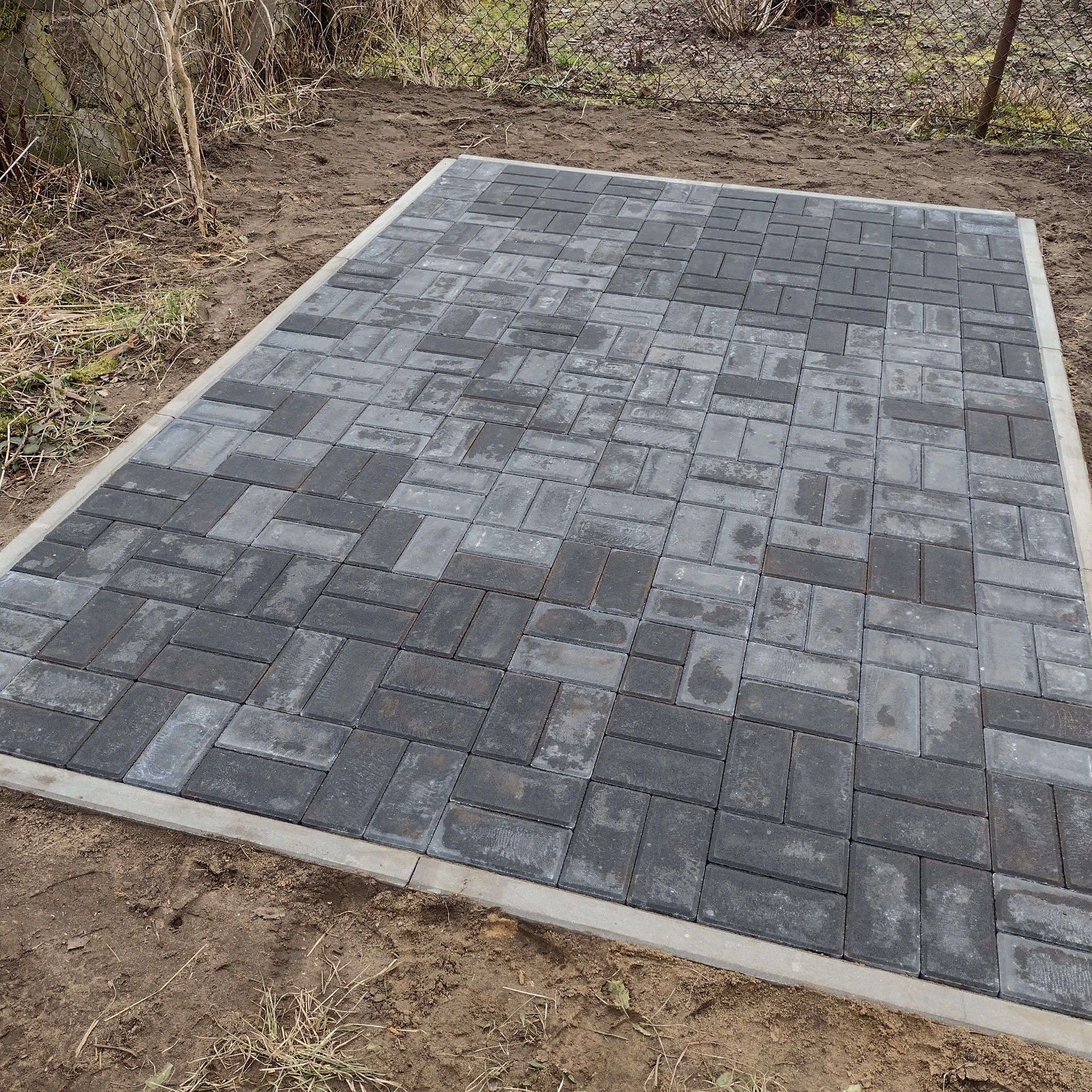 Ułożona, prostokątna powierzchnia z szarej kostki brukowej w wzór jodełki, obramowana betonową obwódką, na tle ziemi i ogrodzenia z siatki.