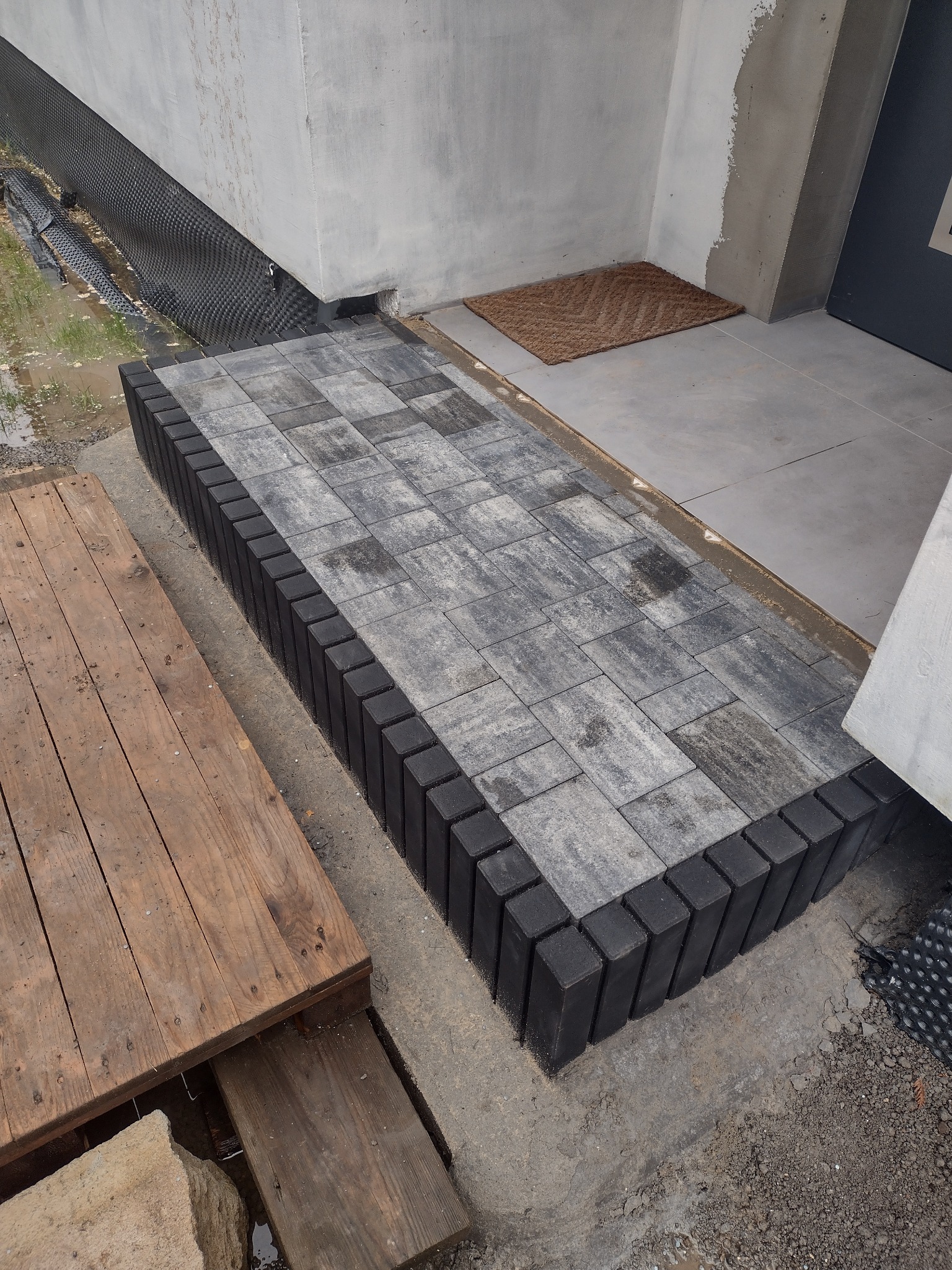 Świeżo ułożona ścieżka z szarej kostki brukowej z czarnym obrzeżem prowadząca do drzwi wejściowych, widoczna mata kokosowa i fragment drewnianego podestu obok.