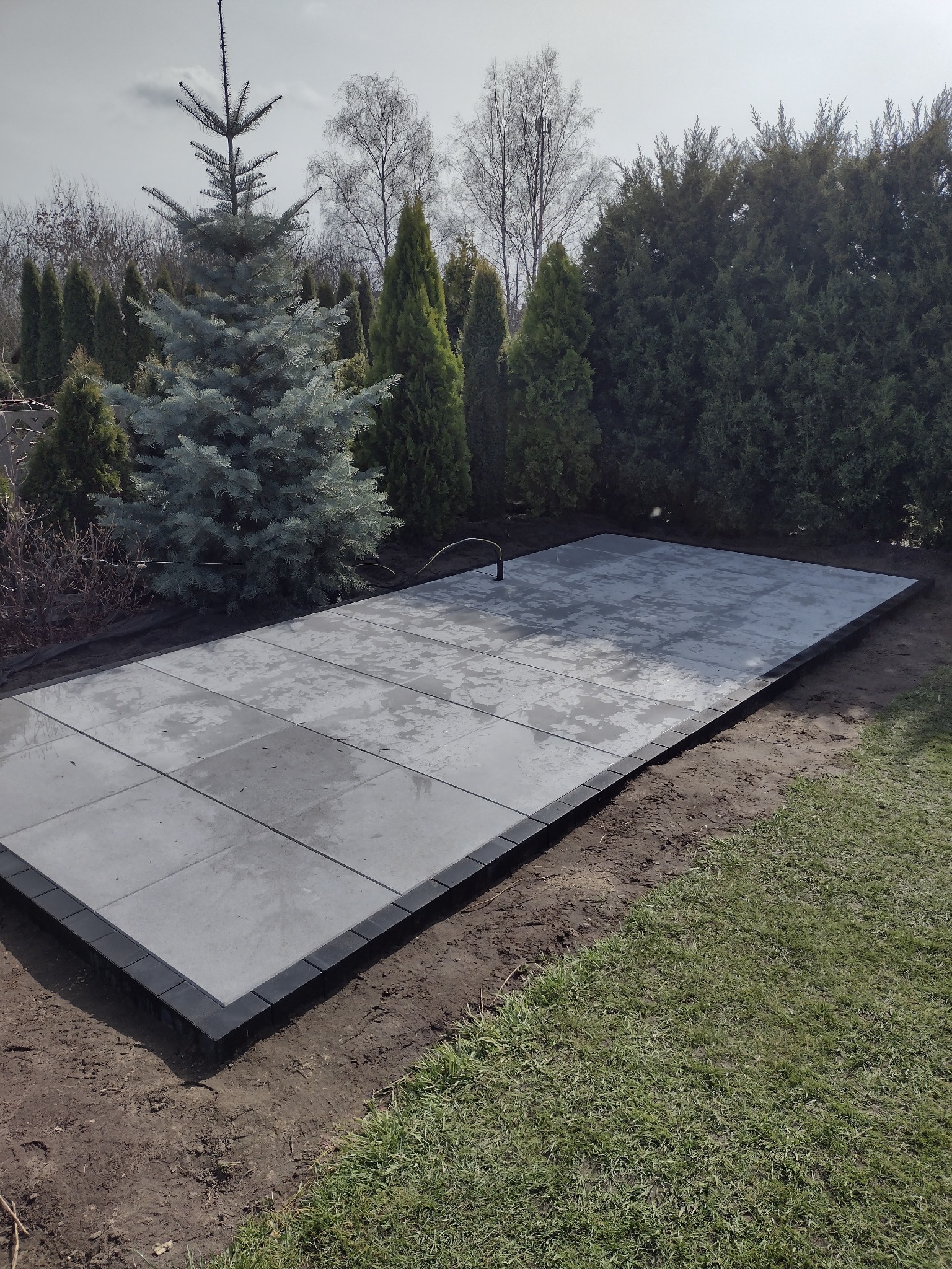Ułożony taras z szarych płyt na gruncie, obramowany ciemnymi kostkami, otoczony zielenią i drzewami iglastymi.