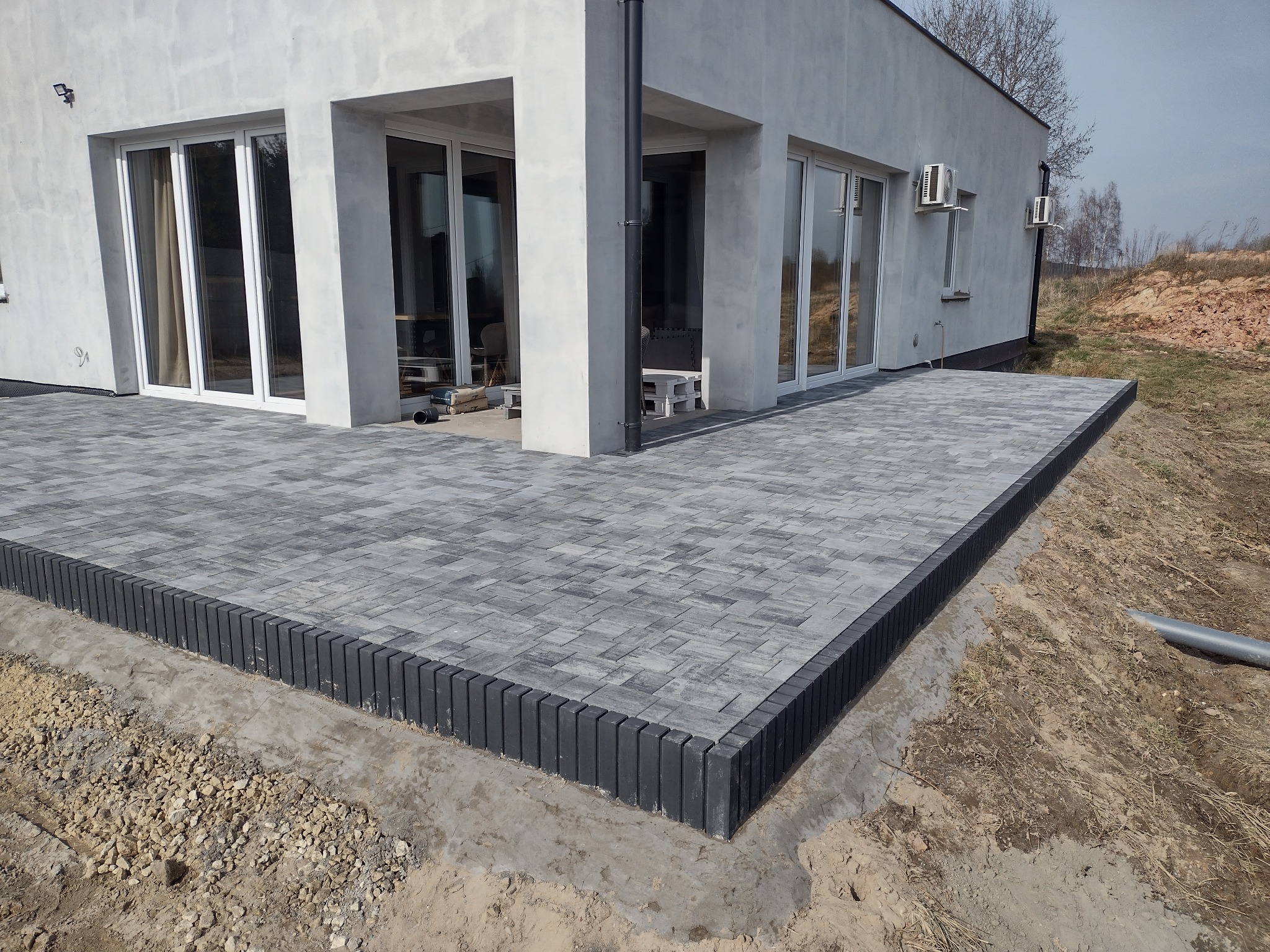 Szara kostka brukowa ułożona jako taras przy nowoczesnym domu z dużymi oknami, widoczne czarne obrzeża i przygotowane podłoże.