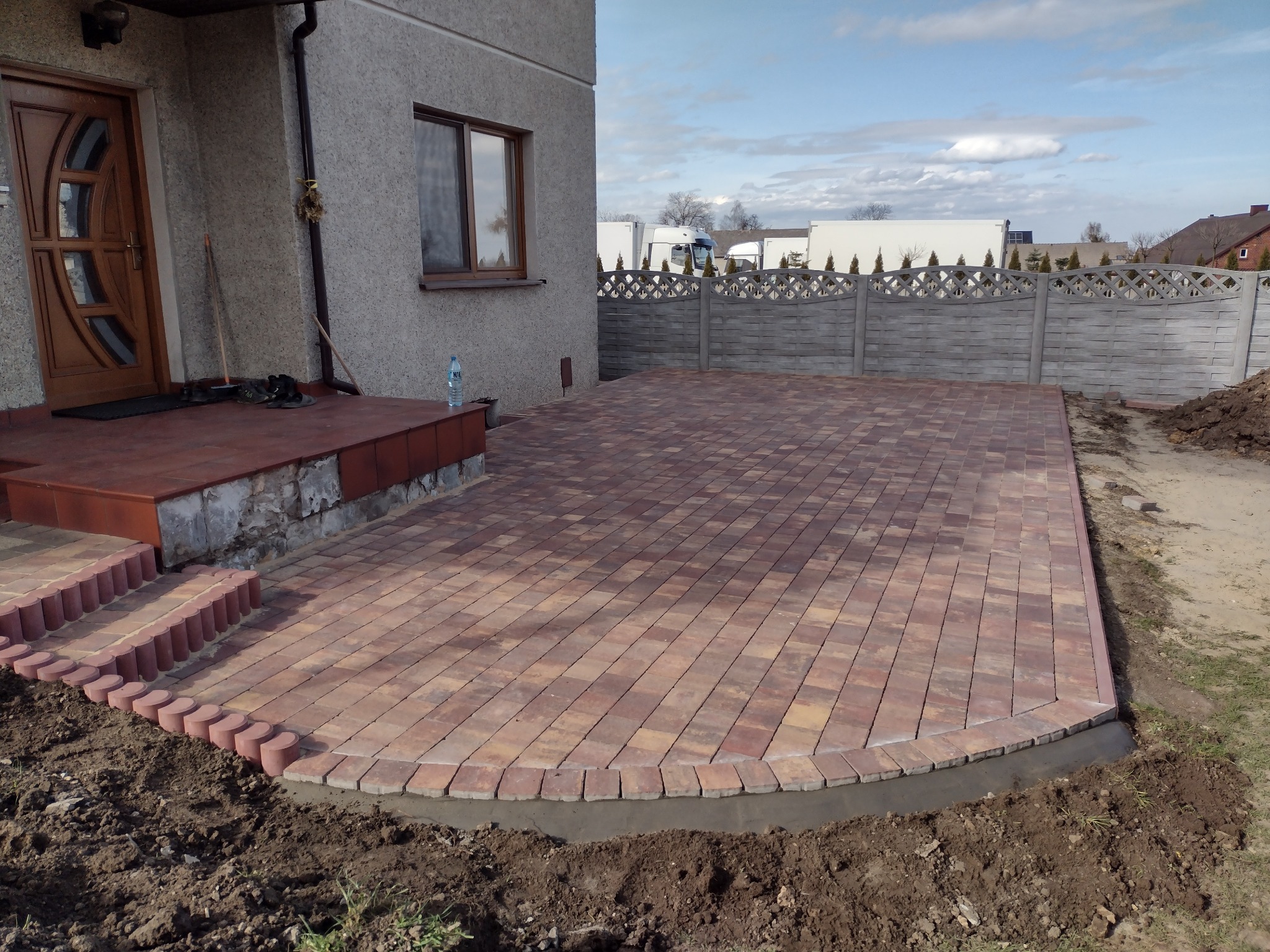 Nowo ułożony, brązowo-pomarańczowy chodnik z kostki brukowej przed domem, z obrzeżem z małych, okrągłych kostek, widoczne schody wejściowe z tego samego materiału i szara betonowa ściana budynku...