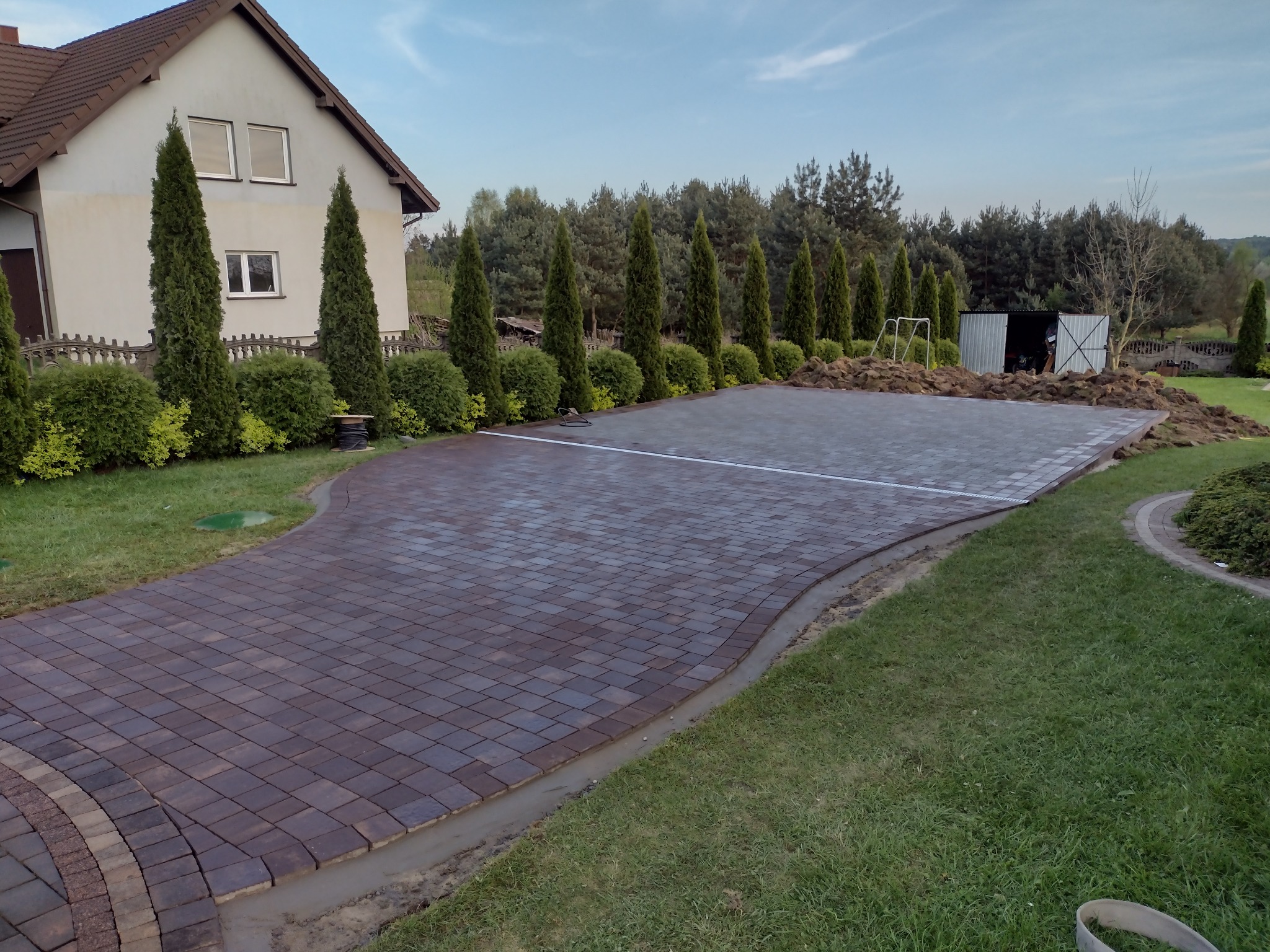 Brązowo-szara kostka brukowa ułożona na podjeździe, z widocznym fragmentem obrzeża i trawnika, w tle dom jednorodzinny i rząd tuj.