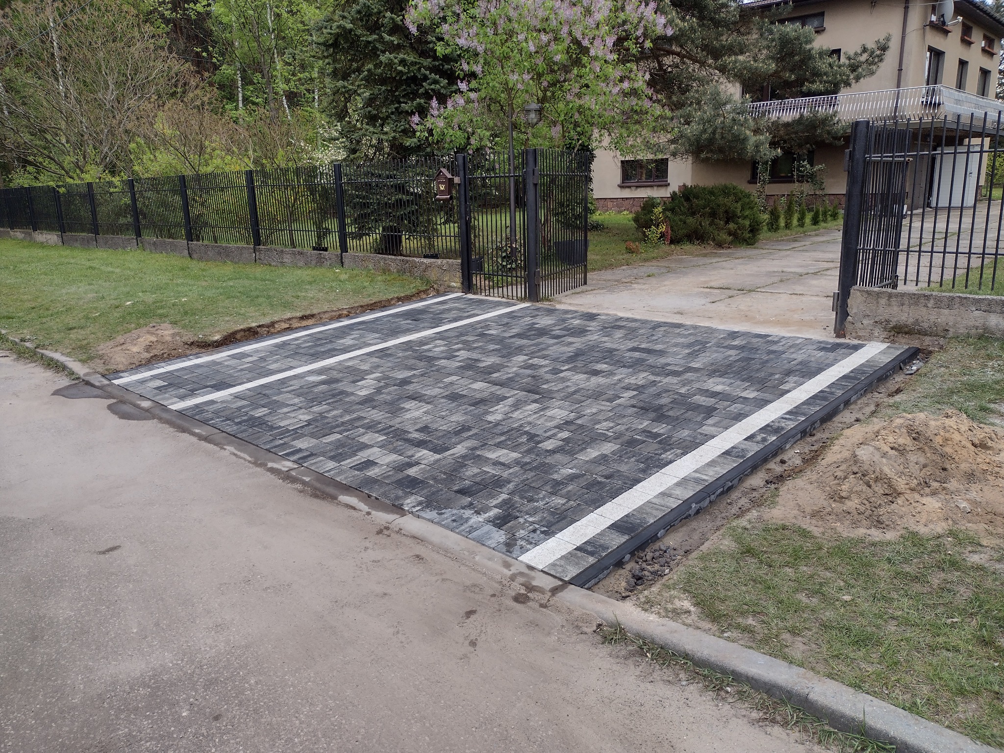 Nowo ułożony podjazd z szaro-czarnej kostki brukowej z białymi liniami, prowadzący do metalowej bramy posesji z domem w tle, widoczny fragment ulicy i trawnika z kupką piasku.