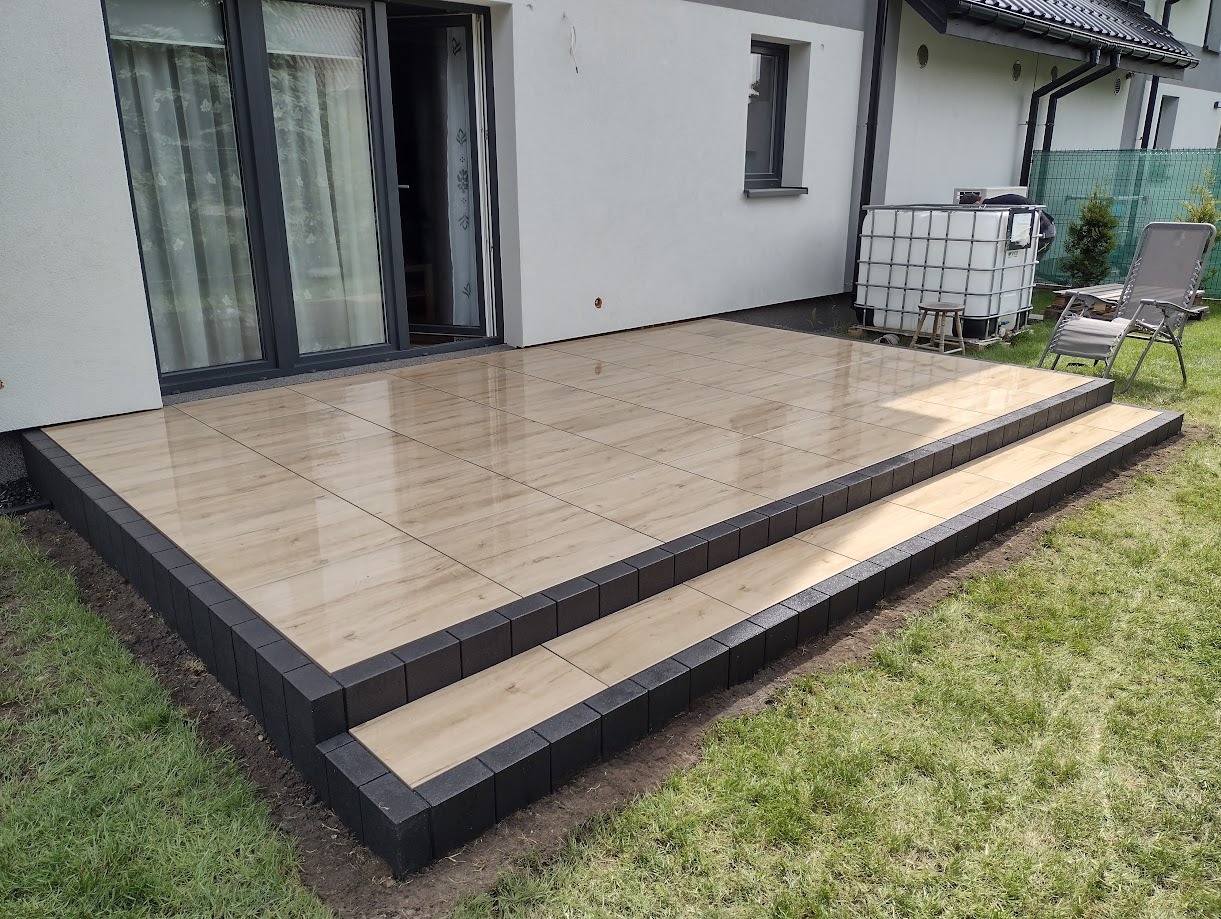 Nowoczesny taras z płytek imitujących drewno, obramowany ciemną kostką, prowadzący do drzwi balkonowych domu z białymi ścianami i zielonym trawnikiem.