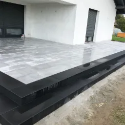 Nowoczesny taras z jasnoszarej kostki brukowej z czarnymi obrzeżami i schodami, widok na biały budynek z szarymi roletami i zielony ogród w tle.