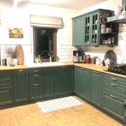 ADREM Adrian Świerzbin Remonty Łazienki Wykończenia - Kuchnia w stylu rustykalnym z zielonymi szafkami, drewnianym blatem i podłogą ułożoną w jodełkę. Nad blatem roboczym widoczne okna z roletami rzymskimi i dekoracje, w tym obrazek i suszone kwiaty.