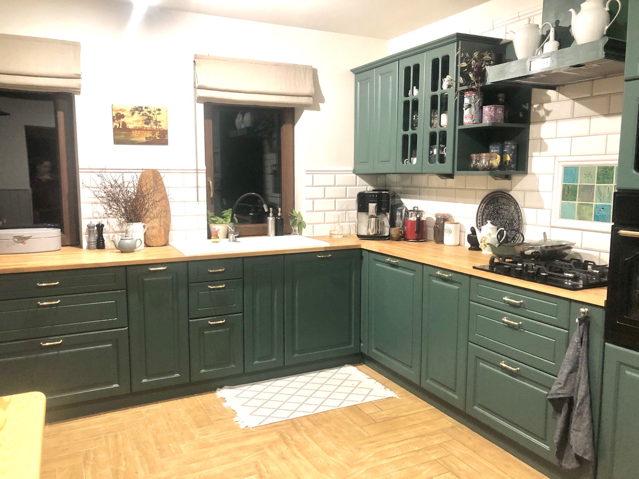 Kuchnia w stylu rustykalnym z zielonymi szafkami, drewnianym blatem i podłogą ułożoną w jodełkę. Nad blatem roboczym widoczne okna z roletami rzymskimi i dekoracje, w tym obrazek i suszone kwiaty.