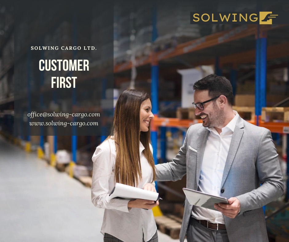 Uśmiechnięta kobieta z dokumentami i mężczyzna z tabletem w magazynie z regałami, logo firmy Solwing Cargo LTD w tle.