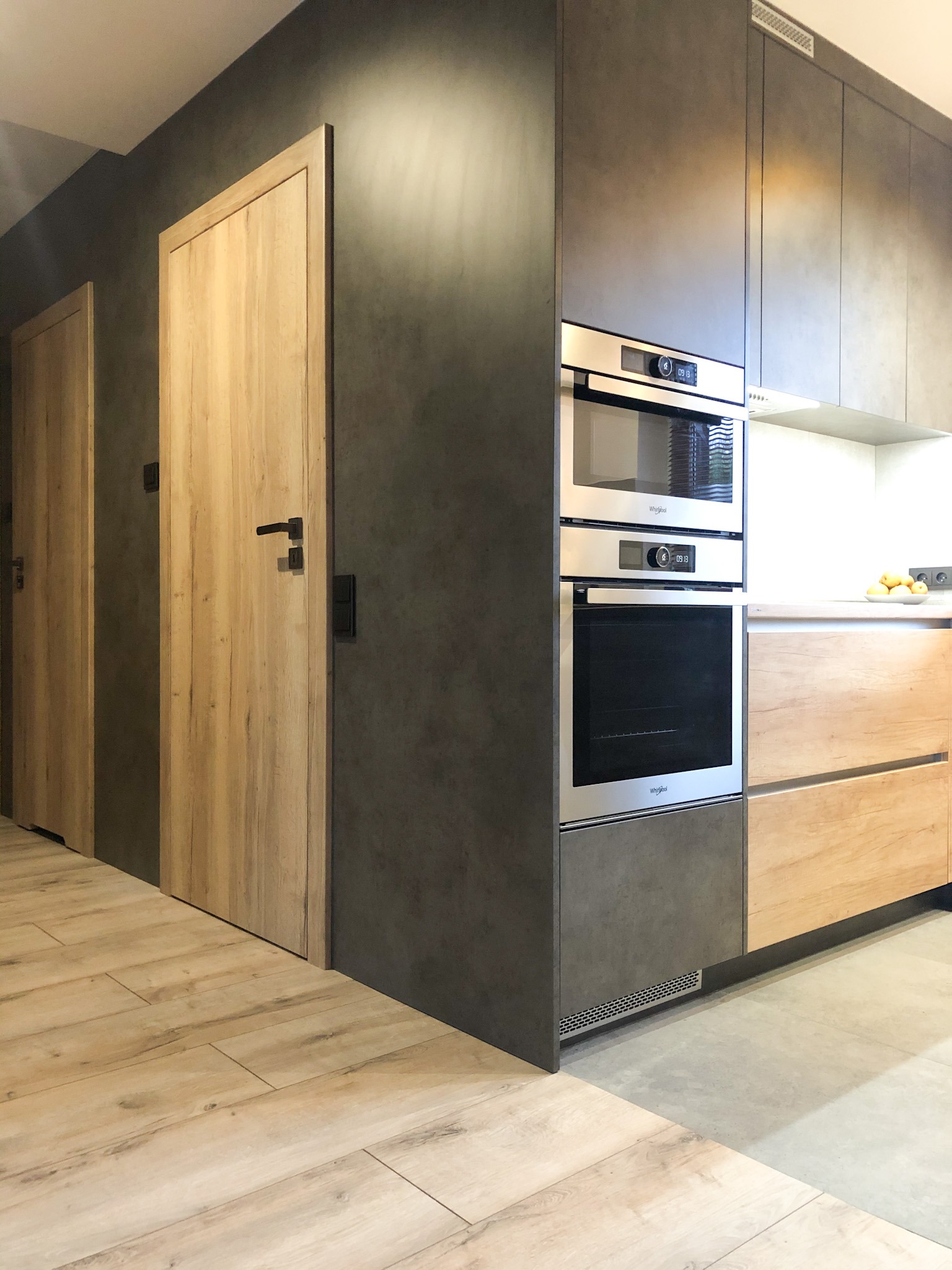 Nowoczesna kuchnia z szarymi szafkami, zabudowanym piekarnikiem i mikrofalówką oraz drewnianymi frontami dolnych szafek, uzupełniona o dwoje drewnianych drzwi z czarnymi klamkami.