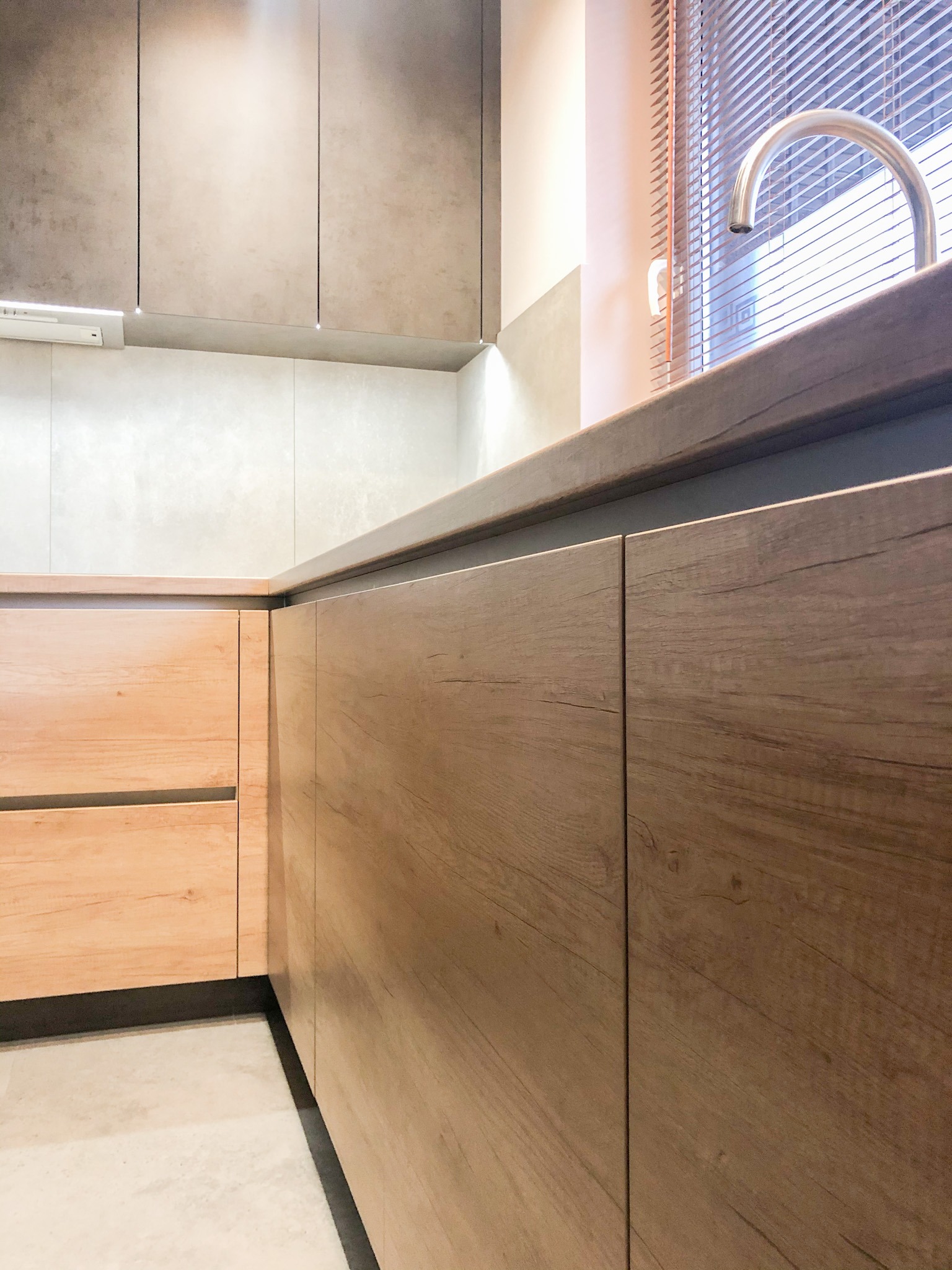 Fragment zabudowy kuchennej z drewnianymi frontami i blatem, widoczny zlew i żaluzje w oknie, minimalistyczny styl.