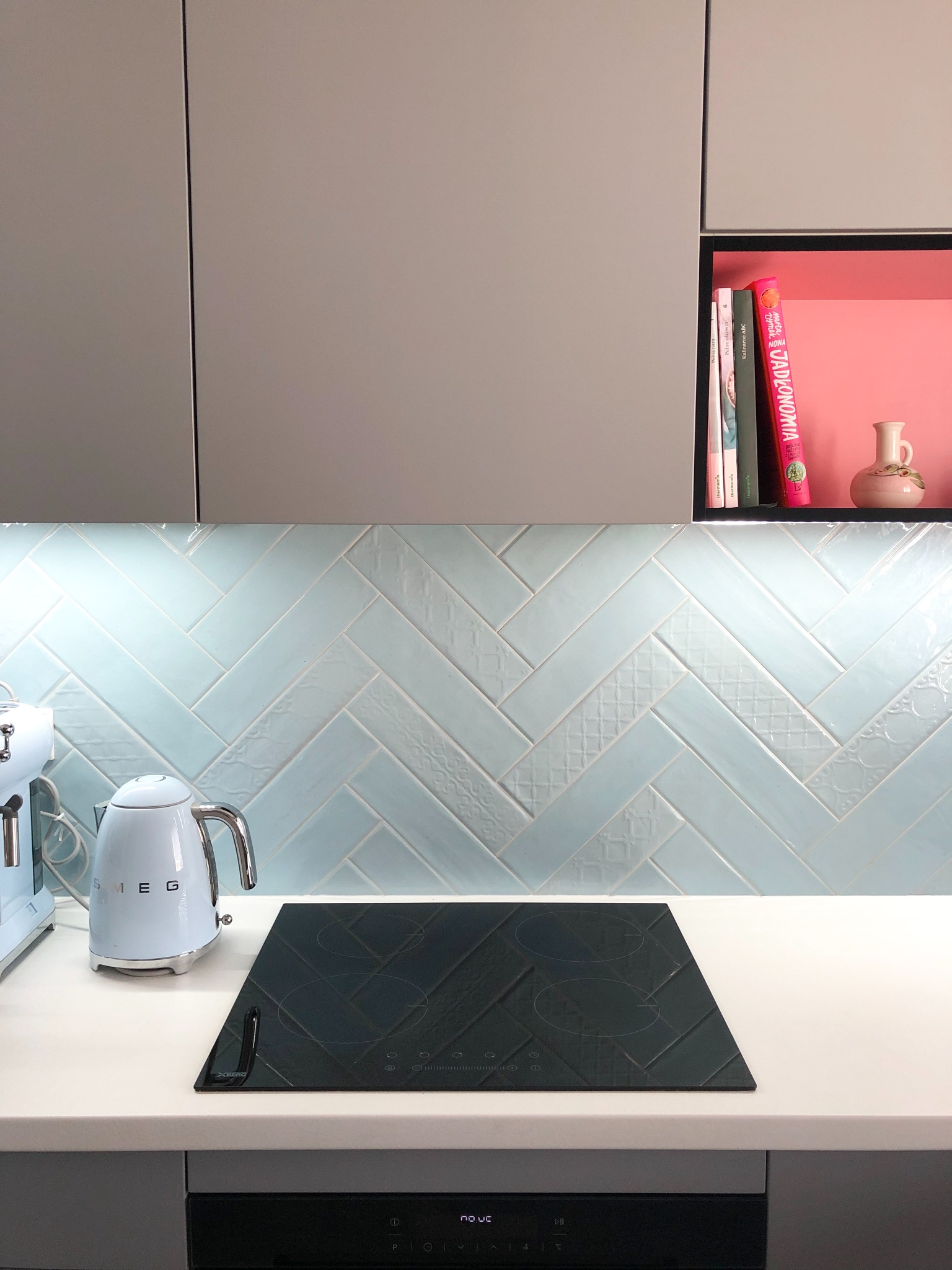 Fragment kuchni z jasnymi, gładkimi szafkami, błękitnymi płytkami ułożonymi w jodełkę z delikatnymi wzorami, czarną płytą indukcyjną i jasnym blatem; widoczny czajnik Smeg i ekspres do kawy...