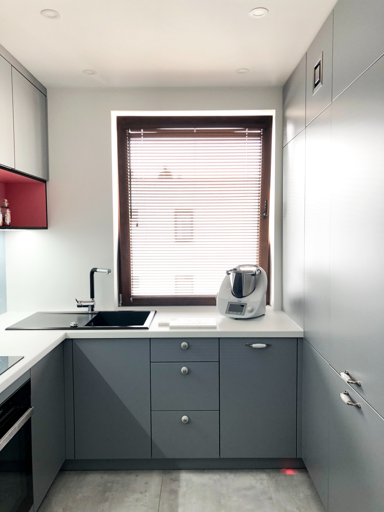 Nowoczesna kuchnia w odcieniach szarości z białym blatem, czarnym zlewem i drewnianymi żaluzjami w oknie. Widoczny robot kuchenny stojący na blacie.