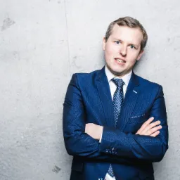 Mężczyzna w niebieskim garniturze i krawacie w kropki, stoi na tle szarej, betonowej ściany z założonymi rękami.