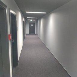 NIERÓWNO POD SUFITEM AGNIESZKA MAJEWSKA - Długi, szary korytarz biurowy z rzędem drzwi po lewej stronie i oświetleniem LED na suficie. Widok perspektywiczny w głąb korytarza.
