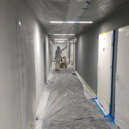 NIERÓWNO POD SUFITEM AGNIESZKA MAJEWSKA - Malowanie natryskowe korytarza biurowego na szaro. Zabezpieczone folią drzwi i podłoga. Pracownik w kombinezonie maluje ściany. Widoczne oświetlenie sufitowe.
