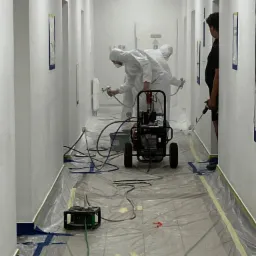 Pracownicy w kombinezonach ochronnych malują ściany w korytarzu biurowym za pomocą agregatu natryskowego. Podłoga zabezpieczona folią malarską.