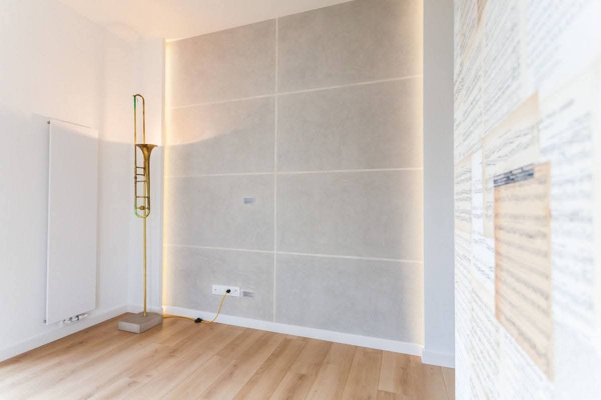 Minimalistyczne wnętrze: drewniana podłoga, ściana z płyt w odcieniach szarości z subtelnym oświetleniem LED, dekoracyjna lampa w kształcie puzonu i ściana z nutami.